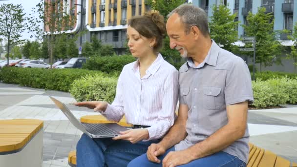 Attraktiv ung kvinde og senior mand bruger laptop sidder på bænken i moderne boligkompleks. Afslappet møde uden for kontoret. En datter underviser i teknologi til sin far – Stock-video