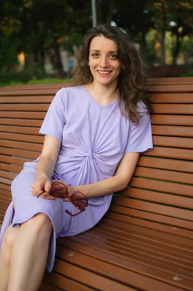 Chica joven en vestido violeta claro está teniendo gran tiempo durante las vacaciones en el verano sentado en el banco, concepto de viaje — Foto de Stock