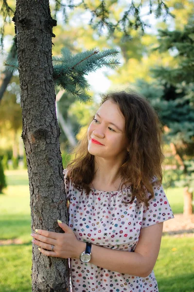 Chica morena joven en vestido está posando sobre el fondo del árbol en el verano en el parque —  Fotos de Stock