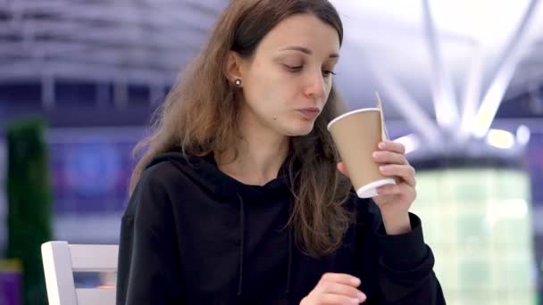 Дівчина тримає чашку кави в руці. Молода дівчина їсть смачний свіжий круасан і п'є деякі напої з паперової чашки в кафе, аеропорту або залізничній станції — стокове відео