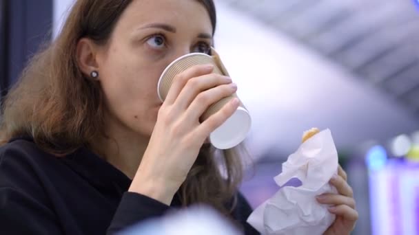 Een meisje met een kop koffie in de hand. Jong meisje is lekker verse croissant eten en drinken wat drank uit een papieren beker in cafe, of luchthaven, of treinstation — Stockvideo