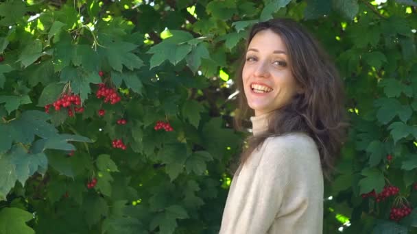 Kırmızı böğürtlen arka planında büyük yeşil ağaçta genç bir kadın, kameraya gülümseyerek poz veriyor.. — Stok video