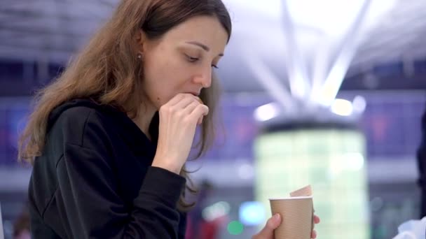 Elinde bir fincan kahve tutan bir kız. Genç kız taze kruvasan yiyor ve kafedeki, havaalanındaki ya da tren istasyonundaki kağıt bardaktan içecek içiyor. — Stok video