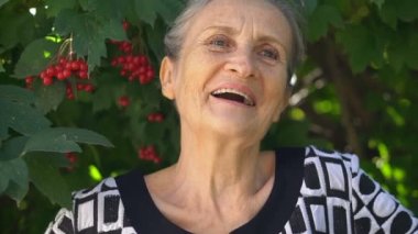 Gri saçlı ve kırışık yüzlü güzel yaşlı büyükanne yeşil ağaç arka planında gülümseme ve mutlulukla kameraya bakıyor. Anneler Günü, mutlu emeklilik.