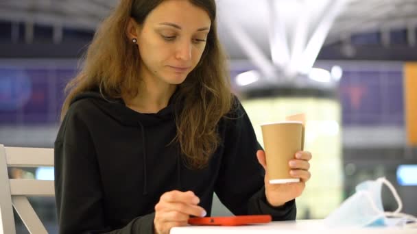 Uma rapariga com uma chávena de café na mão. Menina está comendo croissant fresco saboroso e beber alguma bebida de uma xícara de papel no café, ou aeroporto, ou estação ferroviária — Vídeo de Stock
