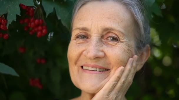 Gyönyörű öreg nagymama ősz hajjal és ráncos arccal néz a kamerába mosollyal és boldogsággal a zöld fa háttér, anyák napja, boldog nyugdíjba vonulás — Stock videók