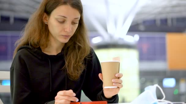 Une fille tenant une tasse de café à la main. La jeune fille mange un délicieux croissant frais et boit une boisson dans une tasse en papier dans un café, un aéroport ou une gare — Video