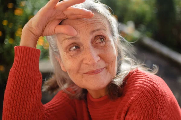 Retrato de mujer mayor con el pelo gris y la cara con arrugas que usan suéter de punto rojo y relajarse en el parque durante el día soleado — Foto de Stock