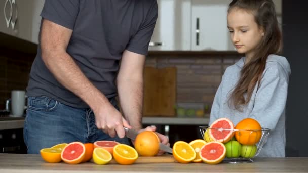 Padre e hija están haciendo jugo fresco de cítricos en casa en la cocina. La niña está lamiendo la mitad de la naranja. Concepto familia, felicidad y estilo de vida. — Vídeos de Stock
