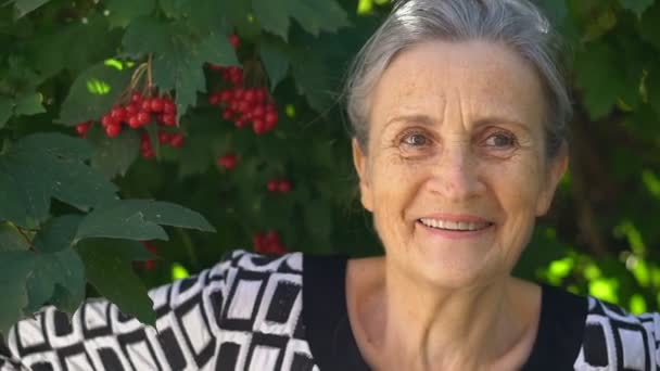 Красива стара бабуся з сірим волоссям і обличчям зі зморшками дивиться на камеру з посмішкою і щастям на фоні зеленого дерева, день матері, щасливий вихід на пенсію — стокове відео