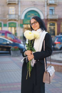 Güzel, orta yaşlı bir kadın bir bahar kasabasında elinde bir buket çiçekle koyu renk bir palto içinde gri saçlı..