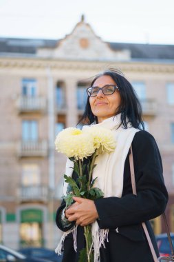 Güzel, orta yaşlı bir kadın bir bahar kasabasında elinde bir buket çiçekle koyu renk bir palto içinde gri saçlı..