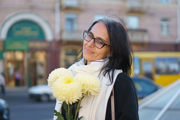 Uma bela mulher de meia-idade ficando grisalho em um casaco escuro em uma cidade de primavera com um buquê de flores. — Fotografia de Stock