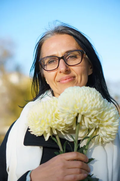 Una hermosa mujer de mediana edad que se pone canosa con un abrigo oscuro en una ciudad de primavera con un ramo de flores. —  Fotos de Stock