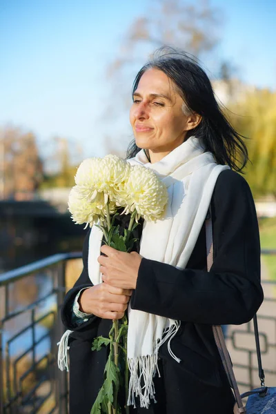 Una hermosa mujer de mediana edad que se pone canosa con un abrigo oscuro en una ciudad de primavera con un ramo de flores. —  Fotos de Stock