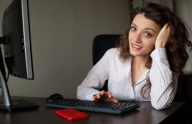 Beyaz gömlekli ve kıvırcık saçlı genç bayan ofis müdürü masada oturuyor ve klavye, rutin iş, serbest çalışma kullanıyor..