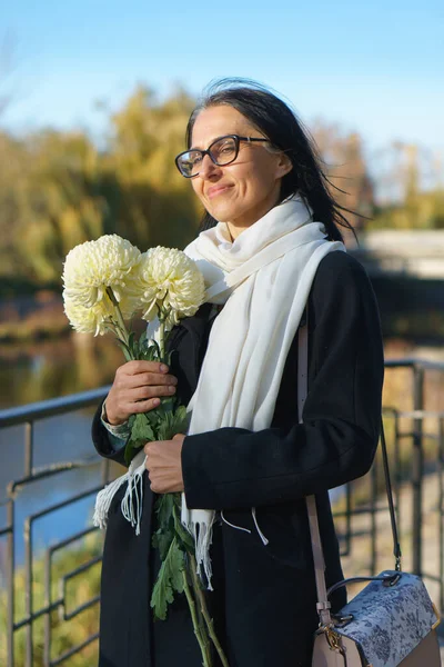 Uma bela mulher de meia-idade ficando grisalho em um casaco escuro em uma cidade de primavera com um buquê de flores. — Fotografia de Stock