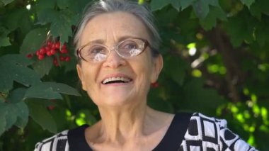 Gri saçlı, kırışık, gözlük takan yaşlı ve güzel bir büyükanne yeşil ağaçta gülümsüyor, anneler günü, mutlu emeklilik.