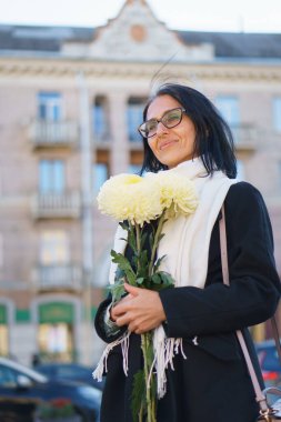 Güzel, orta yaşlı bir kadın bir bahar kasabasında elinde bir buket çiçekle koyu renk bir palto içinde gri saçlı..
