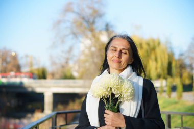 Güzel, orta yaşlı bir kadın bir bahar kasabasında elinde bir buket çiçekle koyu renk bir palto içinde gri saçlı..