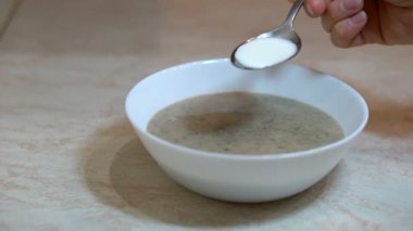 Mutfakta mantar püresi çorbası hazırlama, gümüş metal kaşık kullanarak beyaz kaseye ekşi krema ekleme süreci, sağlıklı yemek, beslenme konsepti.