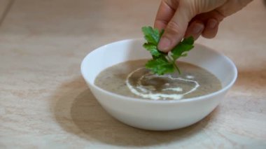 Preparation of mushroom puree soup at the kitchen, process of adding green parsley leaf to white bowl by female hands, healthy eating, nutrition concept