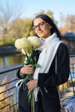 Güzel, orta yaşlı bir kadın bir bahar kasabasında elinde bir buket çiçekle koyu renk bir palto içinde gri saçlı..
