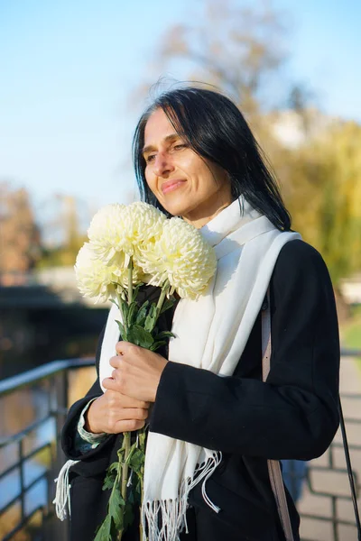 Uma bela mulher de meia-idade ficando grisalho em um casaco escuro em uma cidade de primavera com um buquê de flores. — Fotografia de Stock