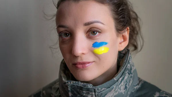 Inomhus porträtt av ung flicka med blå och gul ukrainska flagga på kinden bär militär uniform, obligatorisk värnplikt i Ukraina, jämställdhet begrepp Stockfoto