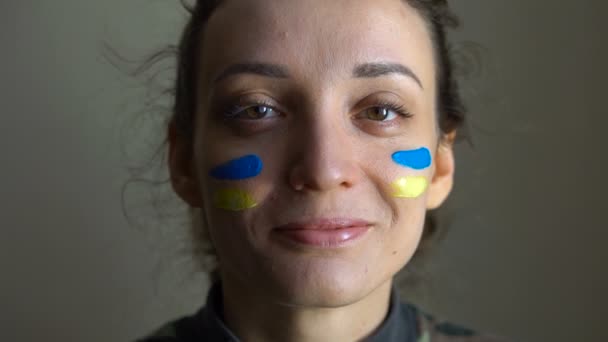 青いと黄色のウクライナの旗を持つ若い女の子の屋内肖像画彼女の頬に軍の制服を着て,ウクライナで強制徴兵,平等の概念 — ストック動画