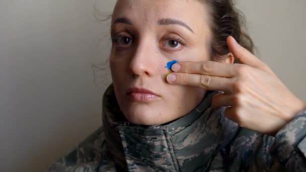 Молода дівчина болить синім і жовтим прапором на щоці, використовуючи пальці у військовій формі, обов'язковому призові в Україні, концепції рівності. — стокове відео