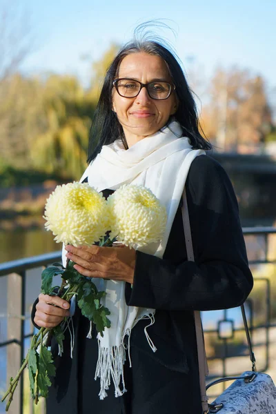 Een mooie vrouw van middelbare leeftijd krijgt grijs haar in een donkere jas in een voorjaarsstad met een boeket bloemen. — Stockfoto