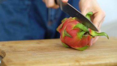 Dişi eller bir ejderha meyvesi veya pitaya 'yı pembe derili ve siyah tohumlu beyaz hamuru masa üzerindeki tahta kesim tahtasıyla kesiyor. Egzotik meyveler, sağlıklı beslenme konsepti