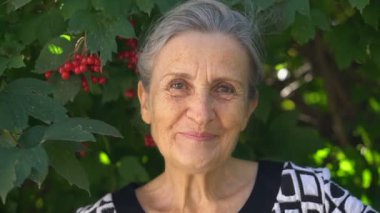 Gri saçlı ve kırışık yüzlü güzel yaşlı büyükanne yeşil ağaç arka planında gülümseme ve mutlulukla kameraya bakıyor. Anneler Günü, mutlu emeklilik.