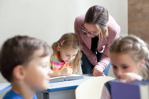 School teacher helping pupils studying, reading book. Lesson for kids in elementary college. Children sitting at desks in classroom. Woman guiding in-person, teaching primary students. Back to school.