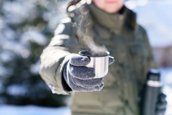 Man giving steamy coffee from thermos. Winter picnic in snowy wonderland. Traveler drinking tea and holding metal cup. Offer hot beverages. Lifestyle moment in nature. Close up of hands in gloves.