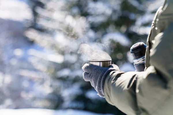 Winter picnic in snowy forest. Man drinking coffee from thermos. Traveler pouring steamy tea and holding metal cup with hot beverage. Lifestyle moment in nature. Close up of hands getting warm.