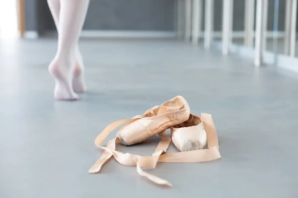 Ballet pointe shoes and ballerina legs in dance studio. Tired girl finishing barre workout and going away barefoot in class room. Close up. Woman has rest and sport break.