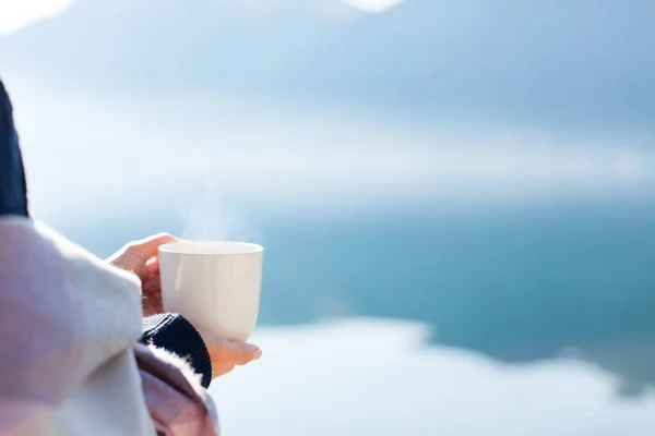 Woman drinking coffee, relaxing at sea beach. Winter picnic with steamy reusable cup by mountains, lake. Girl enjoying ecological travel, slow life, morning tea, peace, calmness. Blue still water.