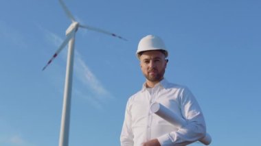 Beyaz kasklı ve klasik tişörtlü bir erkek mühendis elinde projenin büyük yuvarlanmış çizimleriyle rüzgâr türbinlerinin yanında duruyor.