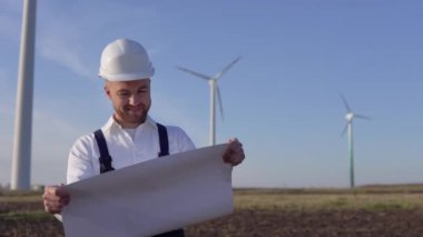 Beyaz kasklı ve klasik gömlekli bir erkek mühendis elektrik santrallerinin kapasitesini inceler ve büyük bir sayfa kağıt üzerinde proje çizimlerini okur.