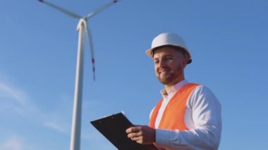 Beyaz kasklı bir erkek mühendis, klasik bir gömlek ve turuncu iş yeleği elektrik santrallerinin tesislerini denetliyor ve çalışan belgelere giriş yapıyor.
