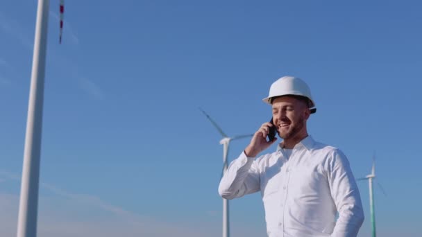 Инженер в деловой одежде и шлеме разговаривает по мобильному телефону на фоне ветряных турбин воздушной электростанции — стоковое видео