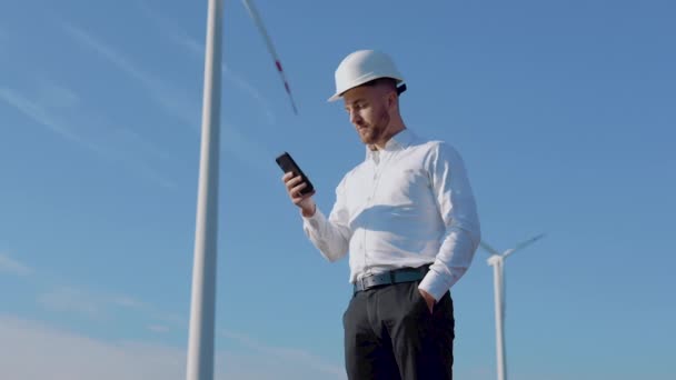 Inženýr v obchodních šatech a helmě pracuje v mobilním telefonu na pozadí větrných turbín letecké elektrárny — Stock video