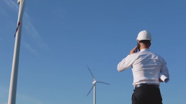 O engenheiro fica de costas para a câmera no fundo dos moinhos de vento e se alegra com as mãos levantadas — Vídeo de Stock