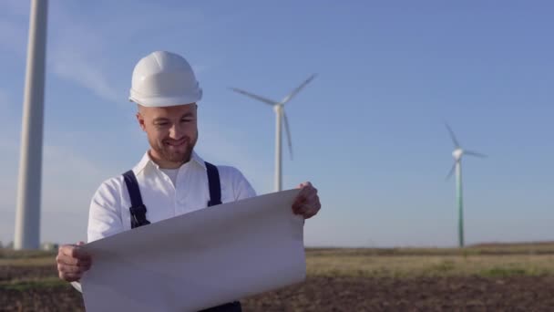 Ένας άνδρας μηχανικός σε ένα λευκό κράνος και ένα κλασικό πουκάμισο επιθεωρεί την ικανότητα των σταθμών παραγωγής ενέργειας και διαβάζει τα σχέδια του έργου σε ένα μεγάλο φύλλο χαρτιού — Αρχείο Βίντεο