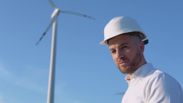 Porträtt av en man i vit hjälm och en klassisk skjorta på bakgrunden av ett vindkraftverk — Stockvideo