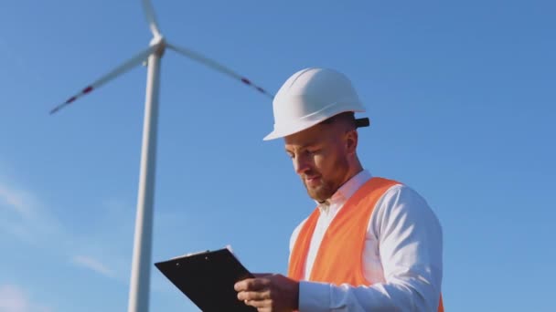 En manlig ingenjör i vit hjälm, klassisk skjorta och orange arbetsväst inspekterar kraftverkets anläggningar och antecknar i arbetsdokument — Stockvideo