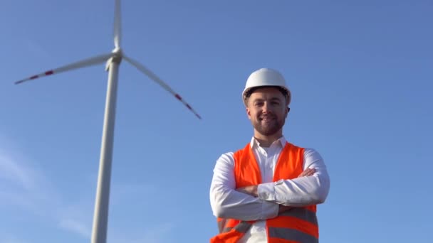 Porträtt av en man i vit hjälm, en klassisk skjorta och en orange arbetsväst på bakgrunden av ett vindkraftverk — Stockvideo