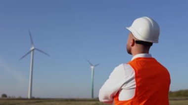 Beyaz kasklı bir erkek mühendis, klasik bir gömlek ve turuncu iş yeleği bir elektrik santralinin rüzgar türbinlerini inceliyor.